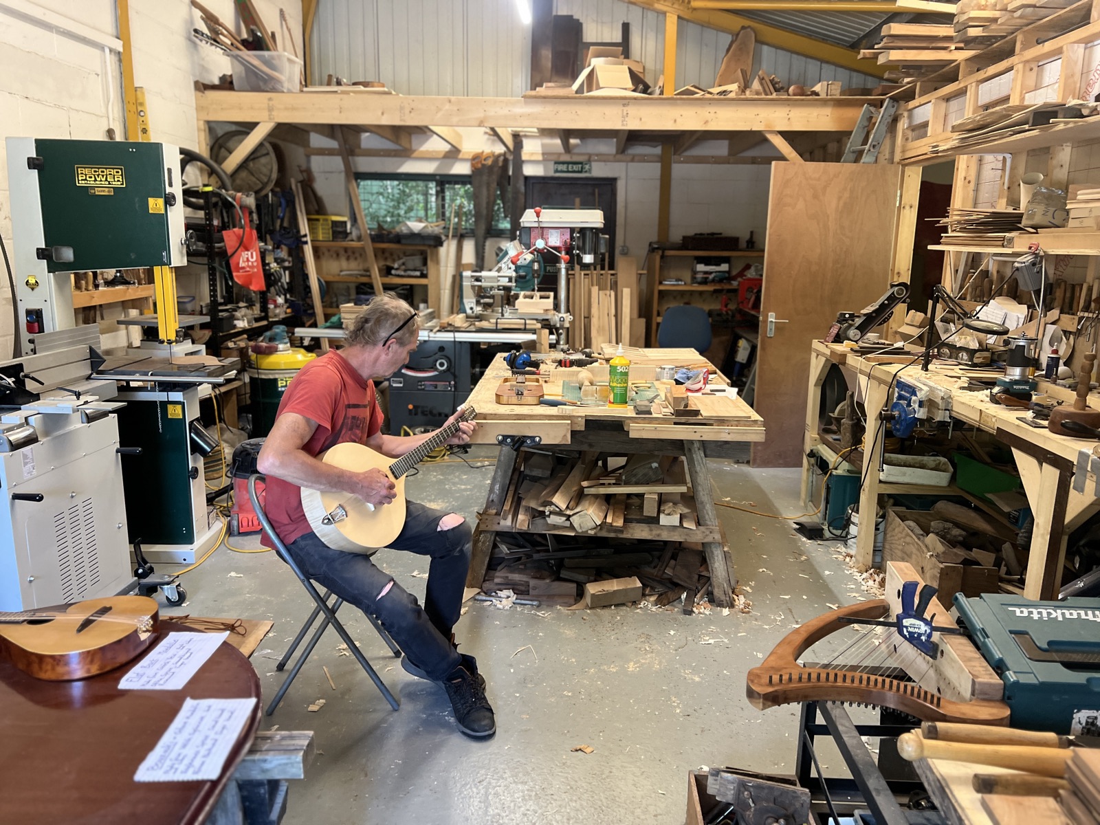 Richard Stephenson playing a bouzouki in his workshop, among the machines, timber and half-made instruments