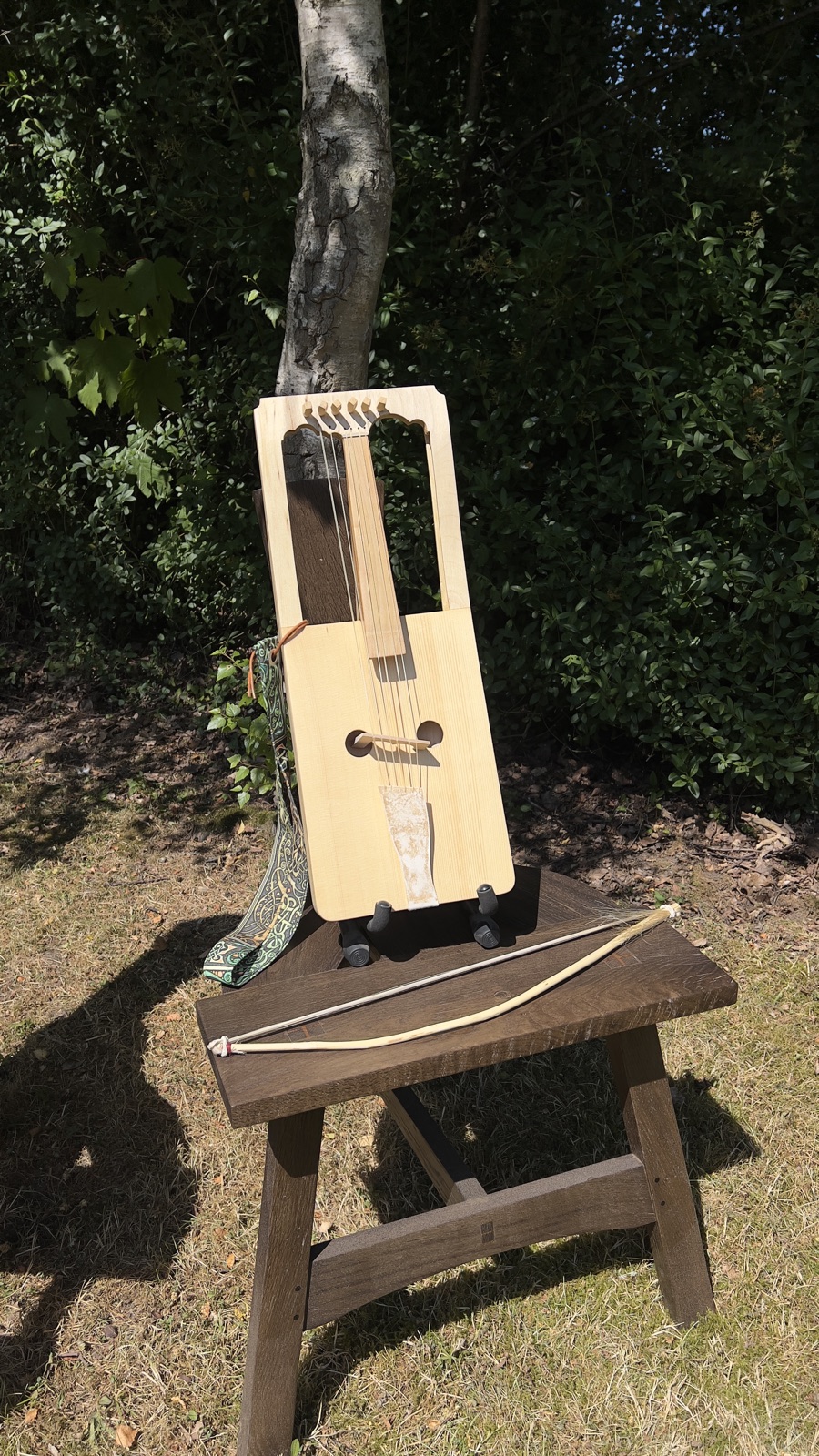 A finished crwth standing on a wooden stool in the garden, its bow beside it